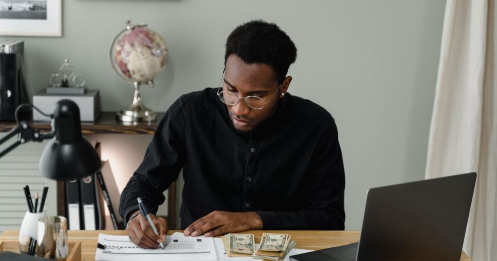 A man at a desk fills in a budget worksheet with a pen, a laptop and a stack of cash beside him