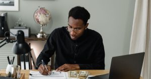 A man at a desk fills in a budget worksheet with a pen, a laptop and a stack of cash beside him