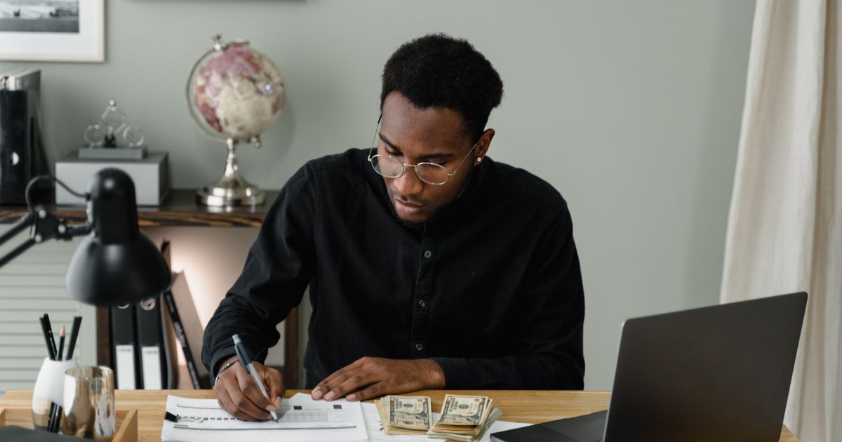 A man at a desk fills in a budget worksheet with a pen, a laptop and a stack of cash beside him
