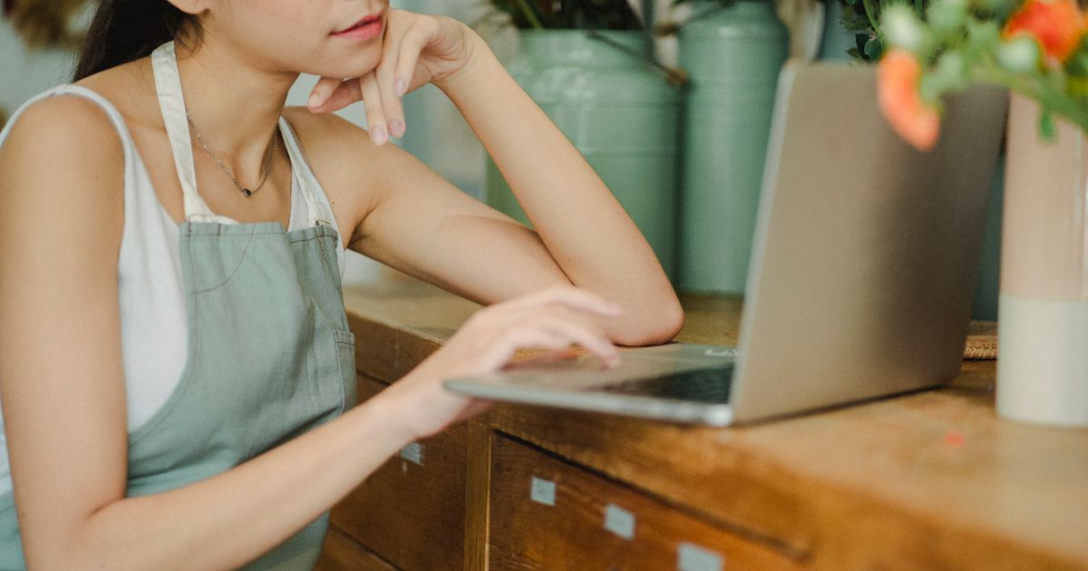 Florist in a green apron resting her chin on her hand while working on a laptop at a wooden shop counter